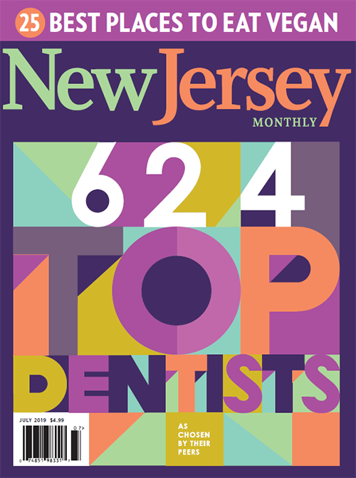 The image features a colorful magazine cover with text that reads  NEW JERSEY  at the top, followed by  25 BEST PLACES TO EAT VEGAN,  below which is a photo of a vibrant dish. The main headline states  624 TOP DENTISTS  with a colorful graphic and a smaller text that says  CHOICE OF THEIR PEERS.  Below the headline, there s an illustration of a top hat with a key and the number  624  prominently displayed. The magazine is from New Jersey Monthly and has a purple border.