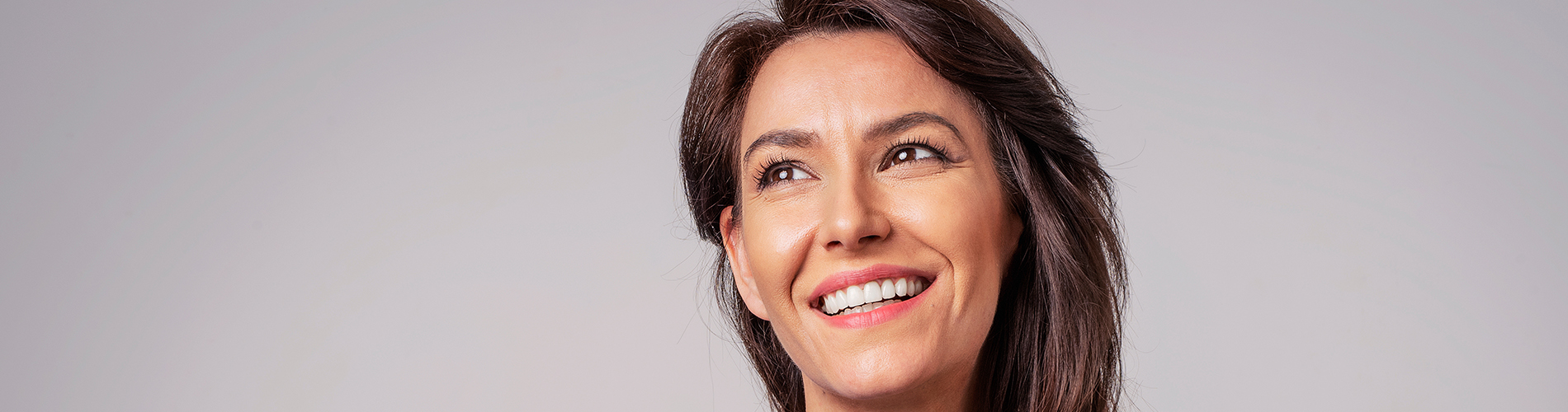 The image is a photograph of a woman with light skin, smiling at the camera. She appears to be in her late twenties or early thirties and has long hair. Her eyes are looking directly at the camera, and she is holding up her index finger near her mouth as if she s making a point or emphasizing something. The background is plain and light-colored, which suggests that this could be a stock photo used for various purposes such as advertising, personal branding, or lifestyle content.