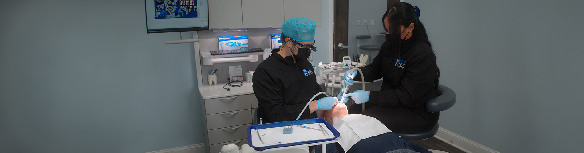 The image shows two individuals in a medical setting, with one person seated at a desk and another standing behind them, both focused on equipment.