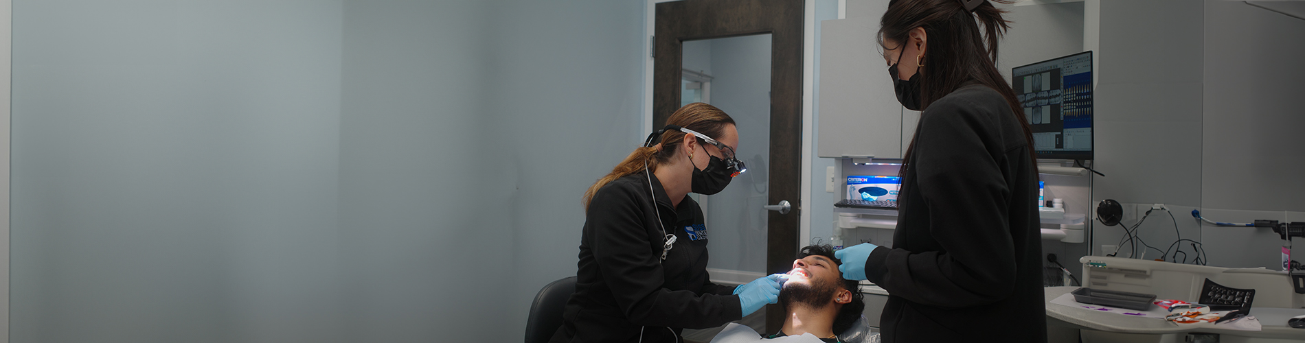 The image shows a panoramic view of an interior space with people, likely a professional setting such as a dental office, where one person appears to be seated at a desk with a computer monitor, while another individual stands nearby, possibly in conversation or observation.
