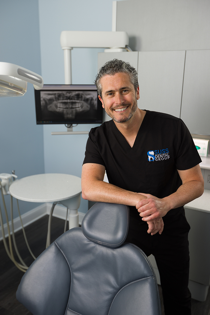 The image shows a man standing in front of a dental chair, smiling at the camera, wearing a black shirt and white scrubs, with a dental office setting behind him.