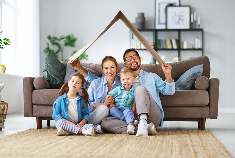 A family of four poses with their hands on a cardboard cutout of a house, smiling at the camera.