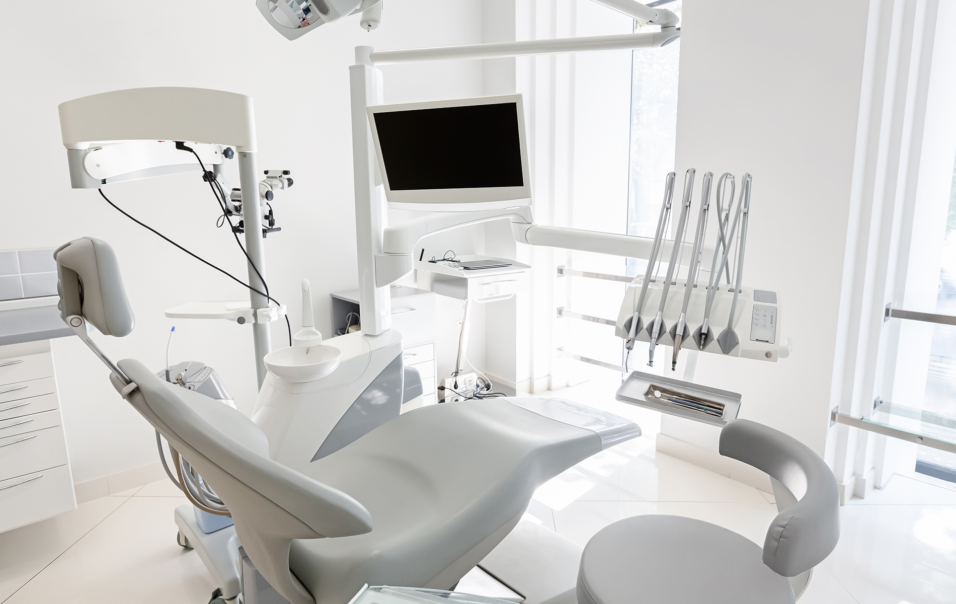 The image shows an interior view of a dental office with modern equipment, featuring a dental chair, examination table, dental tools, and a computer monitor on a desk.