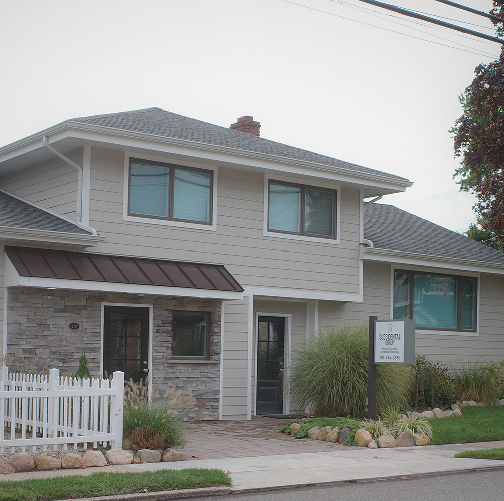 The image shows a two-story residential house with a light beige exterior, a white roof, and a stone facade on the lower level. It features multiple windows, a garage door, and a small front porch. There are landscaped grounds with a well-maintained lawn and garden area in front of the house. The sky is overcast, suggesting an overcast day.