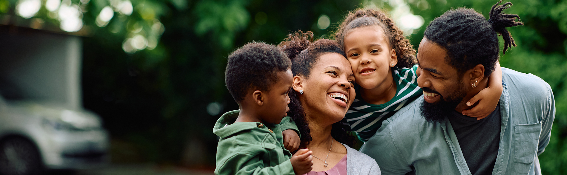The image shows a family of four with two adults and two children, smiling and posing together outdoors during daylight, with one adult holding a child close to them.