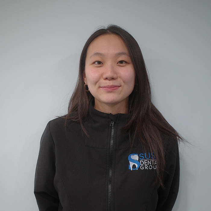 This is a color photograph featuring an individual with long hair wearing a black jacket with a logo on the left side, posing against a plain background.