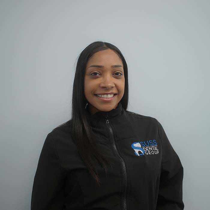 The image shows a person posing for a portrait with a smile, wearing a black jacket with a logo on the left chest area, which appears to be a company name, standing against a light blue background.