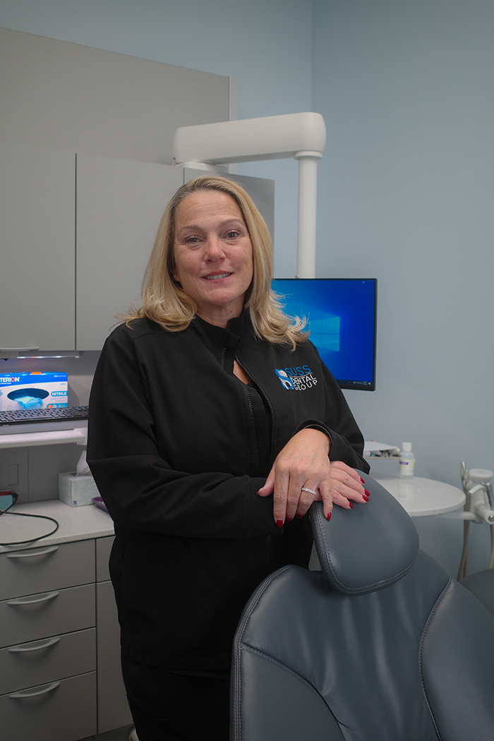 A woman wearing a black jacket stands behind a dental chair in an office setting.