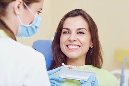 The image shows a woman sitting in a dental chair with a smile, receiving dental care from a professional who appears to be a dentist, with both of them wearing face masks.