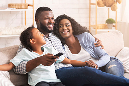 A family of three sitting on a couch, with the father holding a remote control, smiling at the camera.