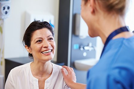 The image shows two individuals in a medical setting  one appears to be a female patient smiling at a male healthcare professional, likely a doctor or nurse, who is standing beside her with a reassuring expression.