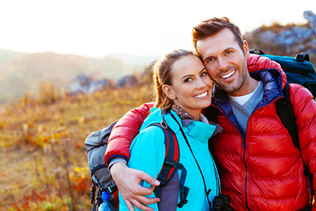 A man and woman are smiling at the camera while posing outdoors with backpacks, suggesting they are on a hiking trip.
