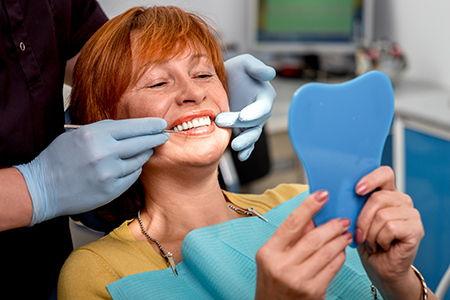 The image shows a woman sitting in a dental chair with a blue mouthguard on her teeth, smiling at the camera while holding it up. There are dental professionals attending to her, one of whom appears to be taking a photograph.