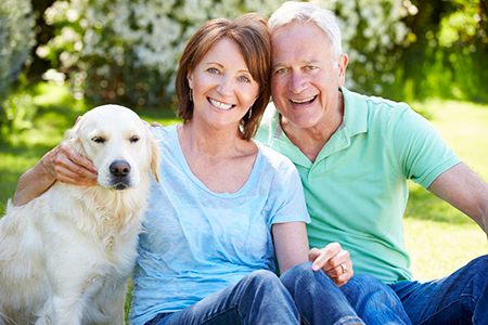 The image shows an elderly couple sitting outdoors with their golden retriever, smiling at the camera.