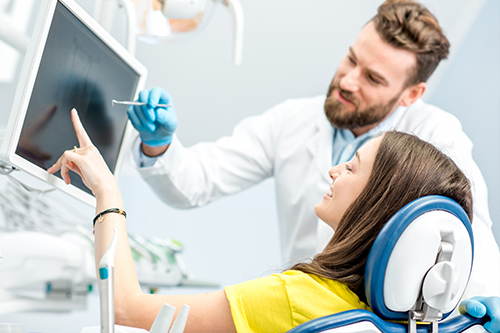 The image shows a dental professional assisting a patient with a computer-aided diagnosis, where the dentist is pointing at an x-ray displayed on a screen while the patient watches attentively.