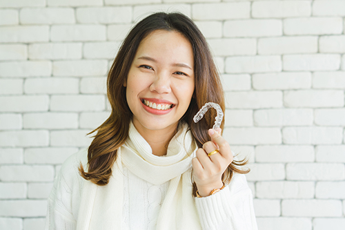A young woman holding a toothbrush with a smile on her face against a brick wall background.