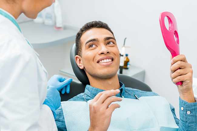 A man in a dental chair receiving oral care from a professional, with a pink object likely used for cleaning teeth held by the patient.