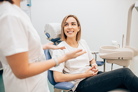 A woman sitting in a dental chair with a smile on her face, being attended by a dental professional who appears to be performing an examination or cleaning her teeth.