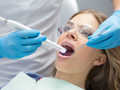 The image depicts a dental professional using a device to examine the teeth of a seated patient, with both individuals wearing protective gloves and masks.
